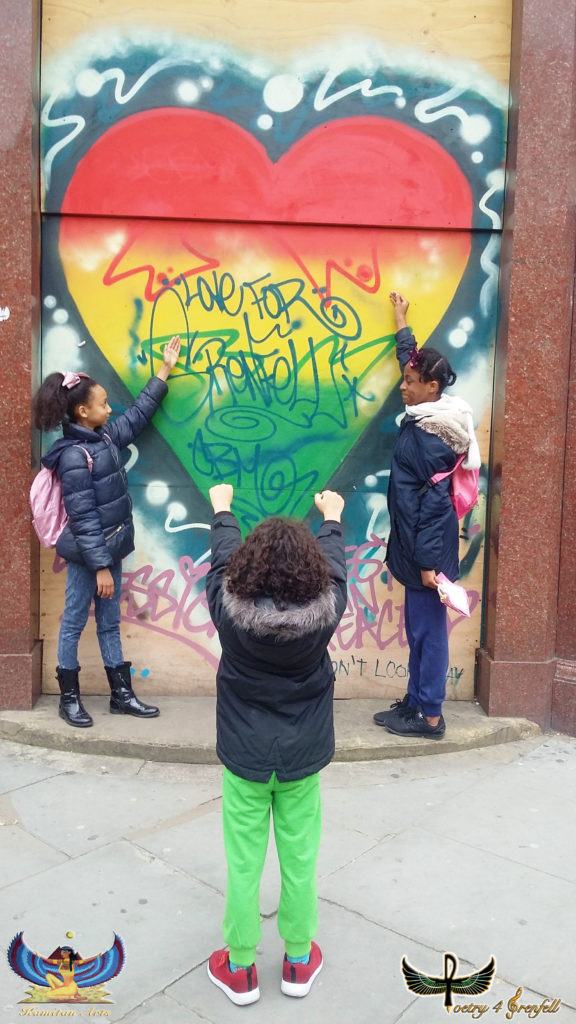 Photograph of three children in front of a red, yellow and green heart mural that says 'Love for Grenfell'.  Two of the children have their hands up touching the wall. 